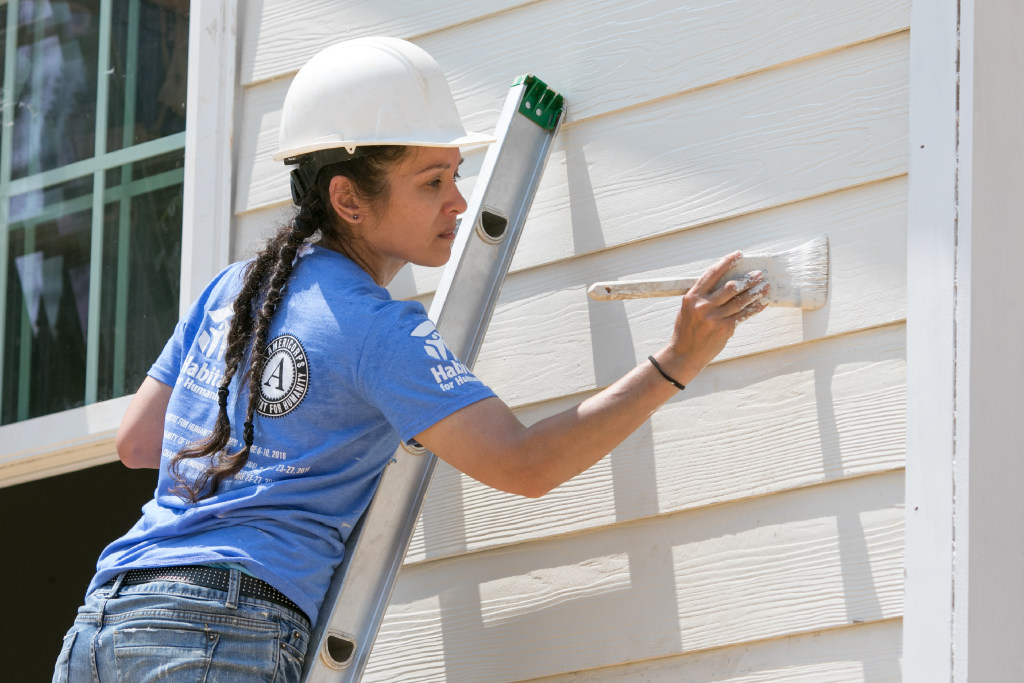 Woman on a ladder wearing a hard hat and reaching over to her right to paint the siding of a house