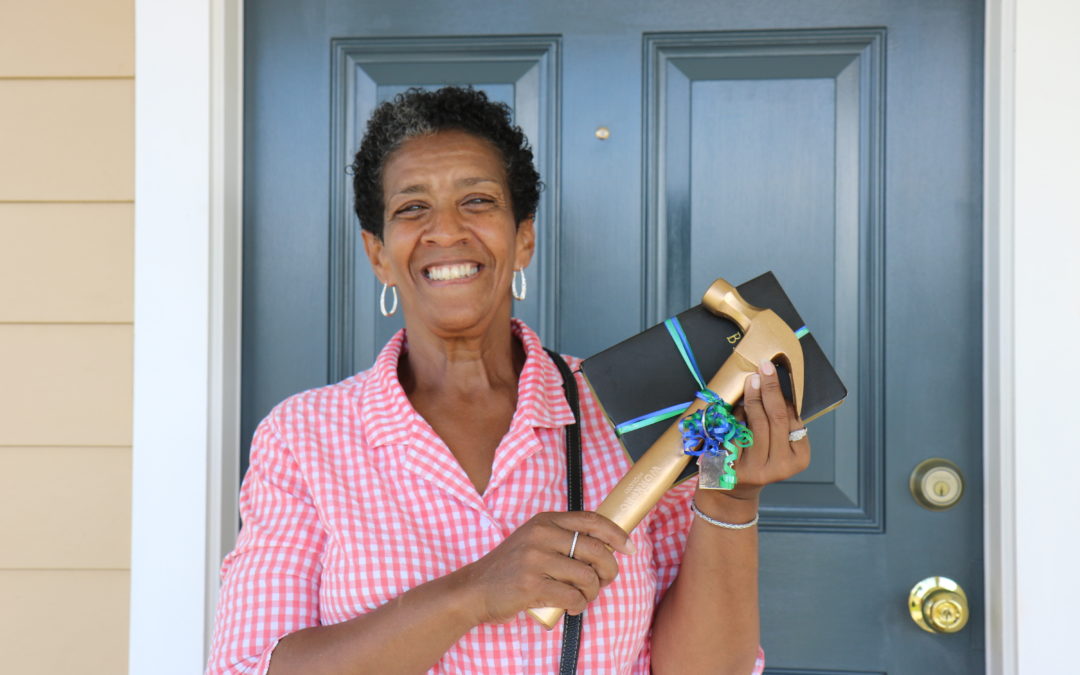 A Habitat homeowner poses in front of a blue door with a gold hammer and a bible.