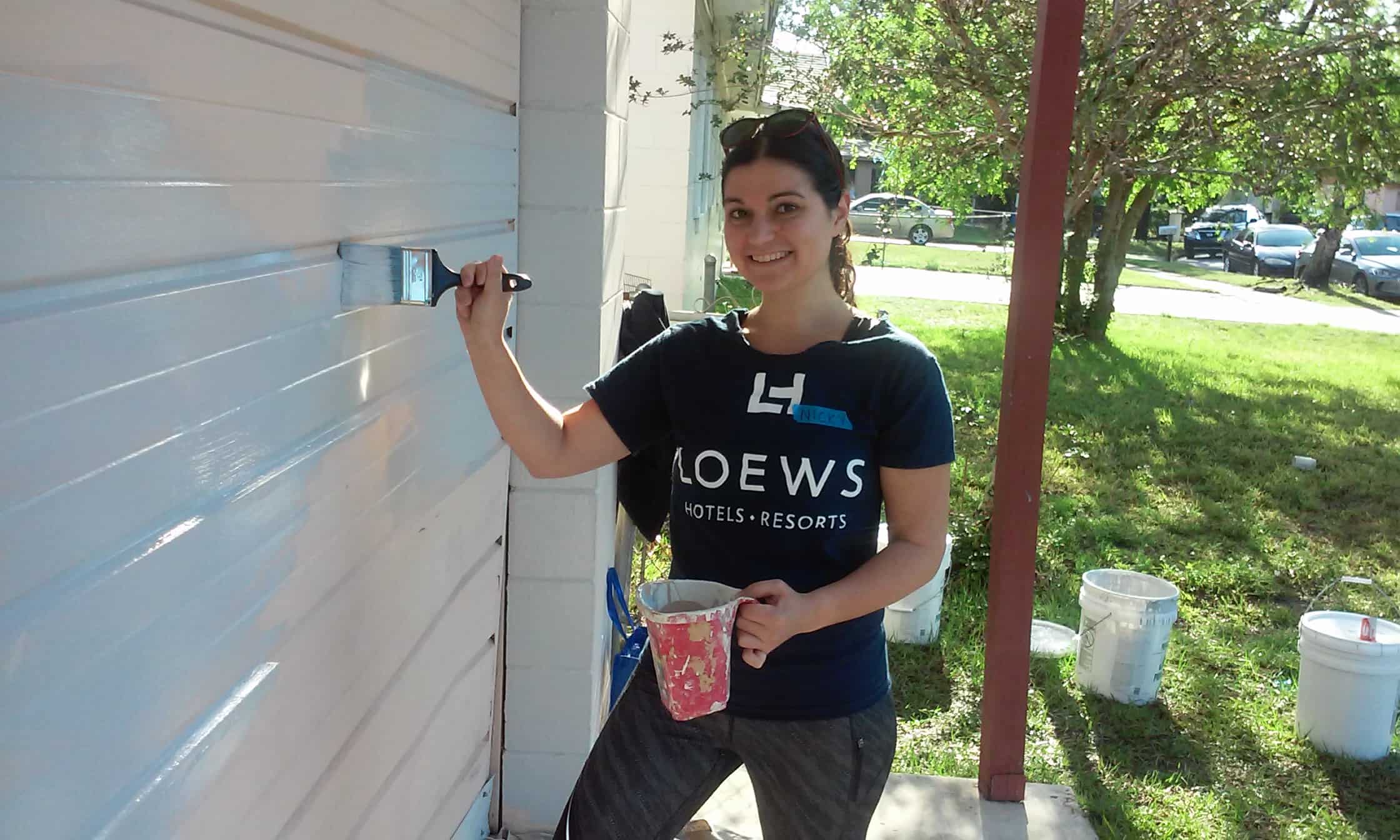 Woman smiling at the camera and painting the side of a house light blue with Neighborhood Revitalization volunteer day