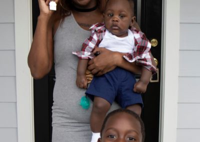 Woman and two young boys smiling and holding house-shaped keychain in front of black front door