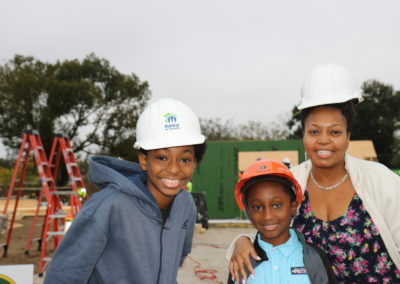 Woman and two boys wearing hard hats and smiling in front of in-progress house