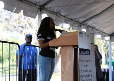 Women standing at microphone on podium with "Lowe's International Women Build Day" sign next to her