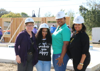 Four women wearing hard hats and smiling in front of in-progress home