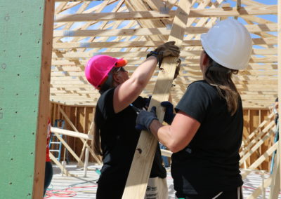 Two women wearing hard hats and raising wooden beam below trusses