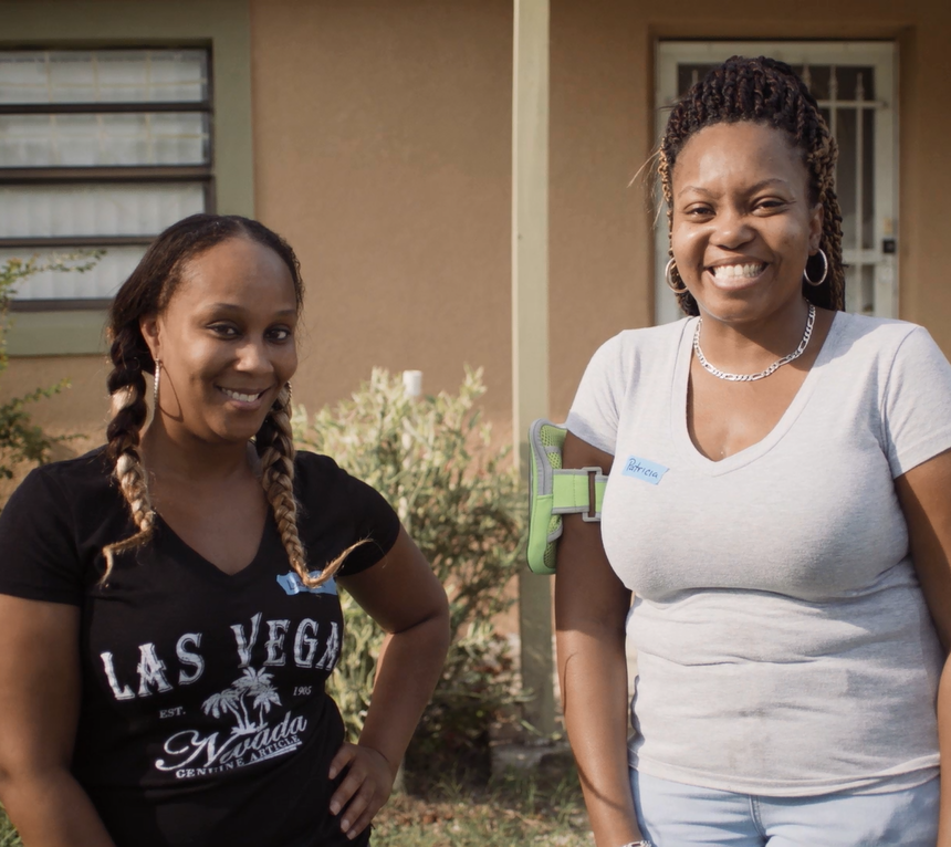 Duas mulheres sorrindo e em pé na frente de uma casa com paisagismo