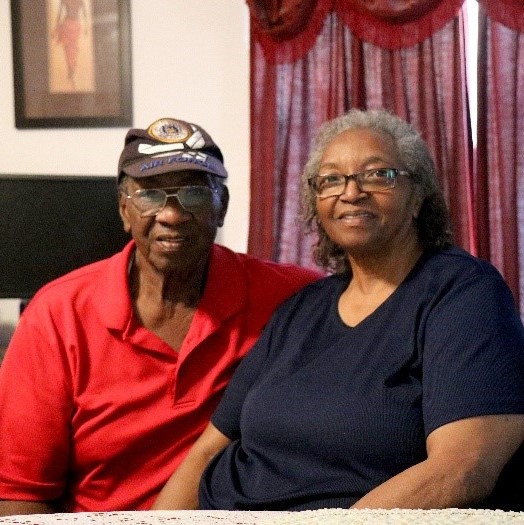 Man and woman smiling and sitting in front of maroon window curtain