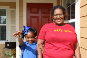 Woman and young girl holding blue house key chain out in front of red door and completed beige house