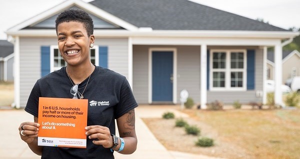 Woman wearing black Habitat for Humanity shirt holding grey paper that says "1 in 6 U.S. households pay half or more of their income on housing. Let's do something about it." in front of completed grey house