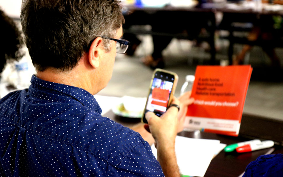 Man wearing polka dot shirt taking a photo on his phone of orange paper with wording on it regarding the affordable housing crisis.