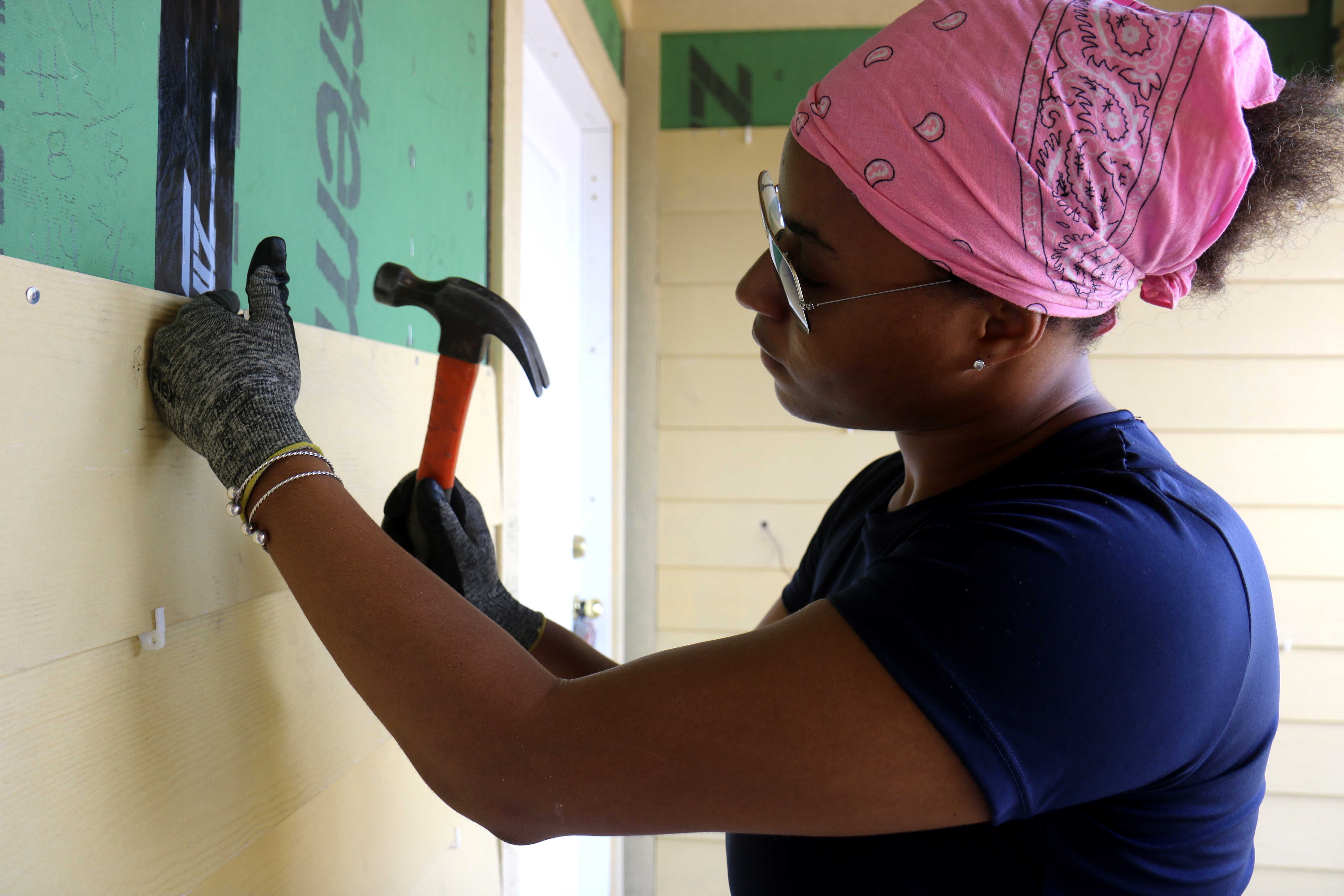 Volunteer wearing pink bandanna holding hammer to install siding on an in-progress house