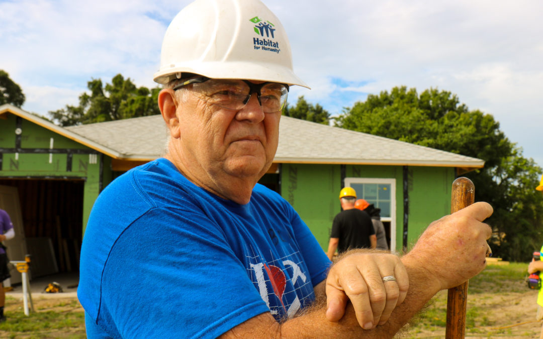 Man wearing white construction hat and protective glasses and holding wooden tool standing in front of partially constructed house