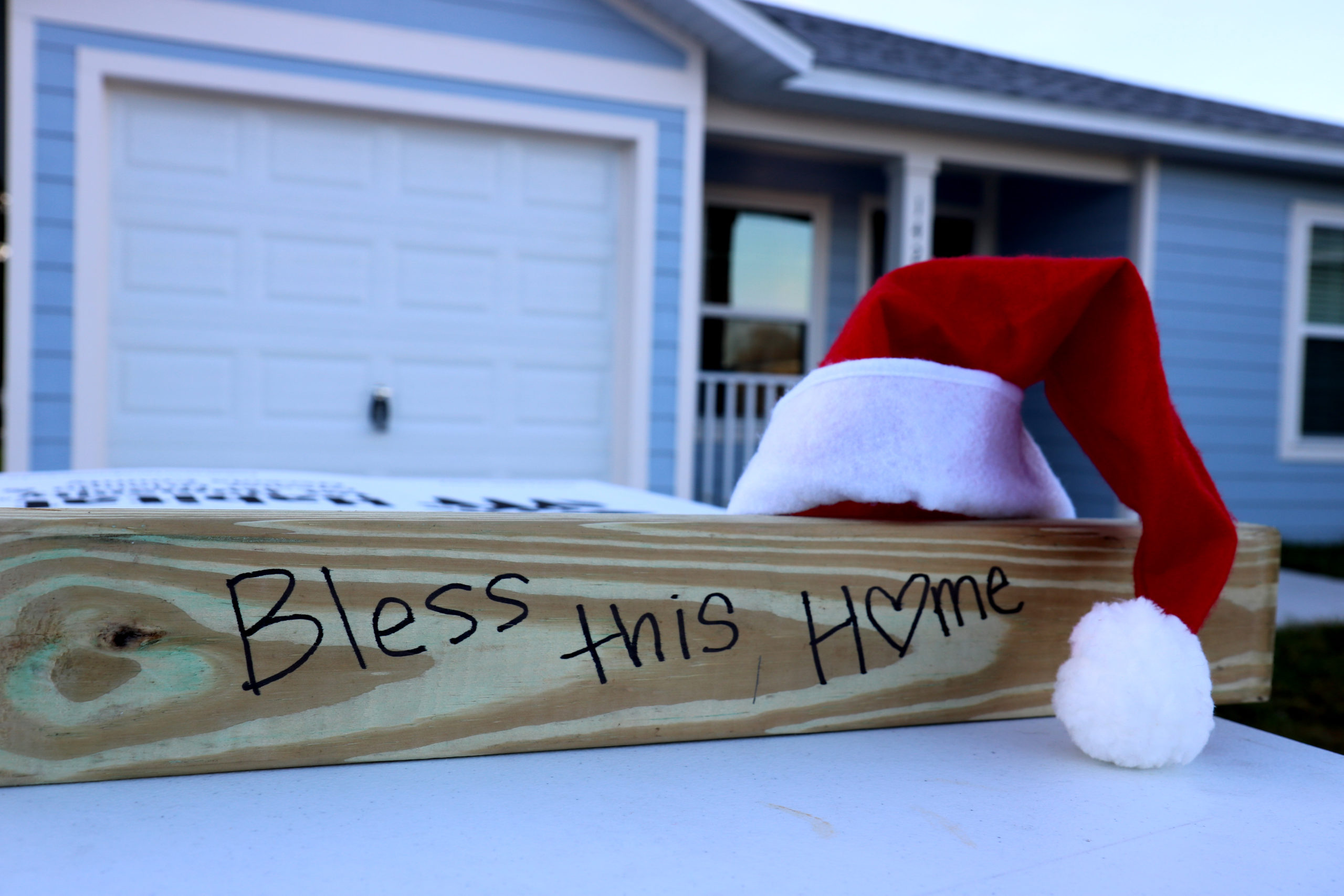 Wood beam reading "Bless this Home" with a Santa hat resting on top, in front of completed house.