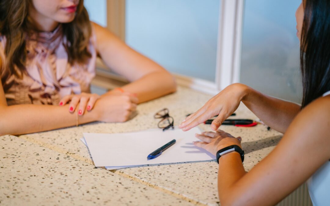 To women talk at a table with a stack of paper, pens, and glasses in between them.