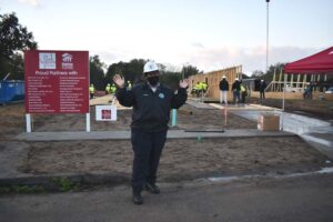 A woman waves with both hands while standing in front of dirt lot