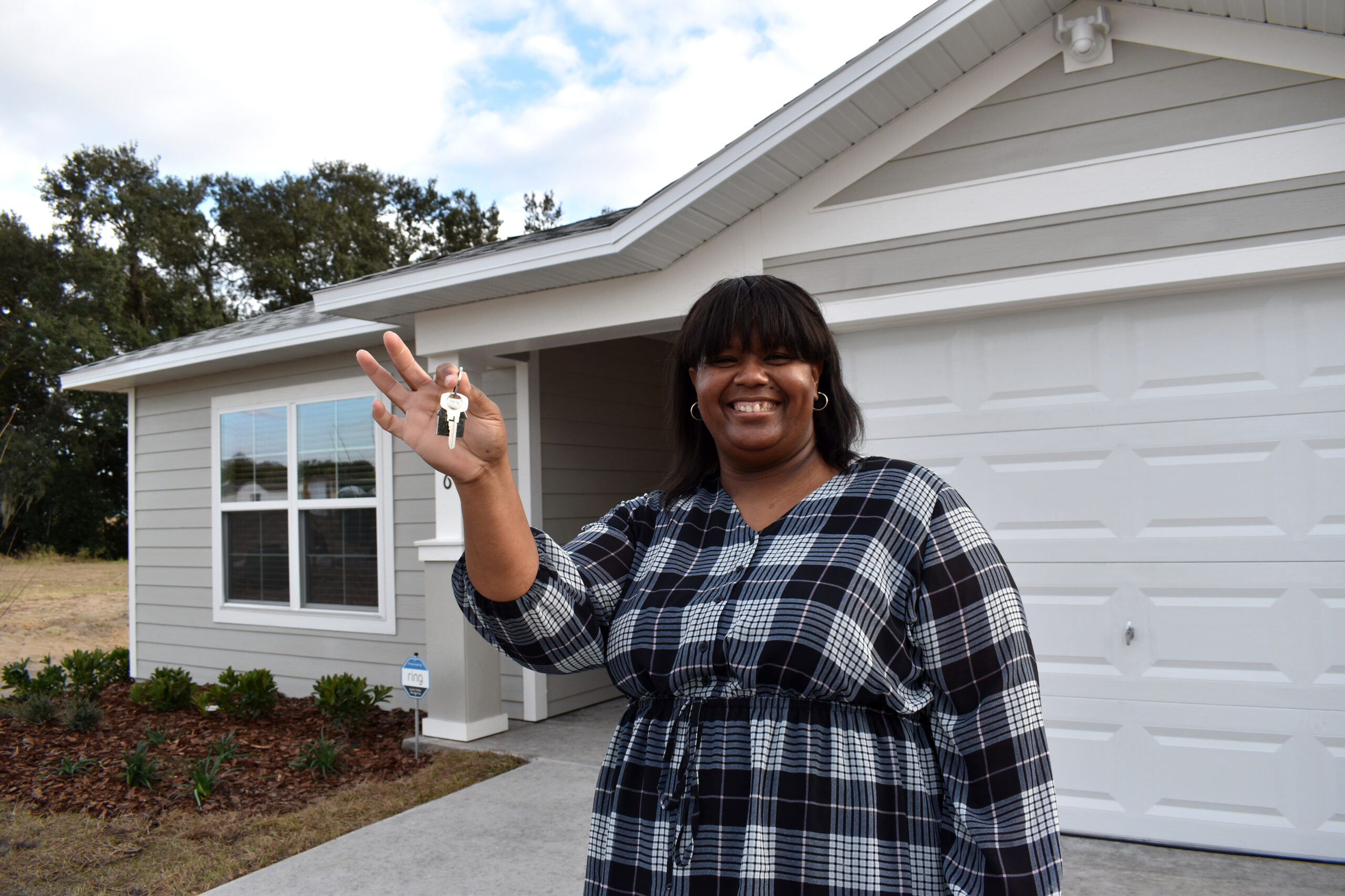 A smiling woman holds up her keys in front of her home.