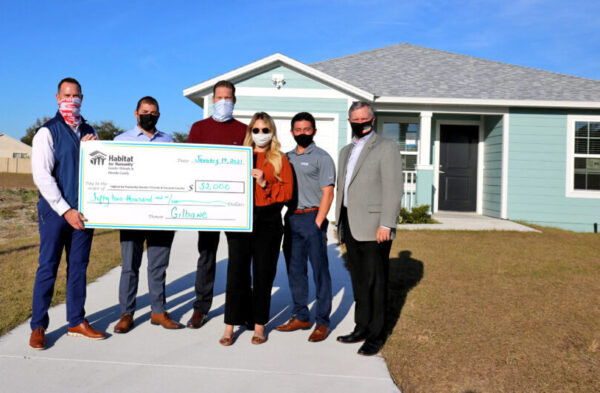 Six people stand in front of a house holding a large check.